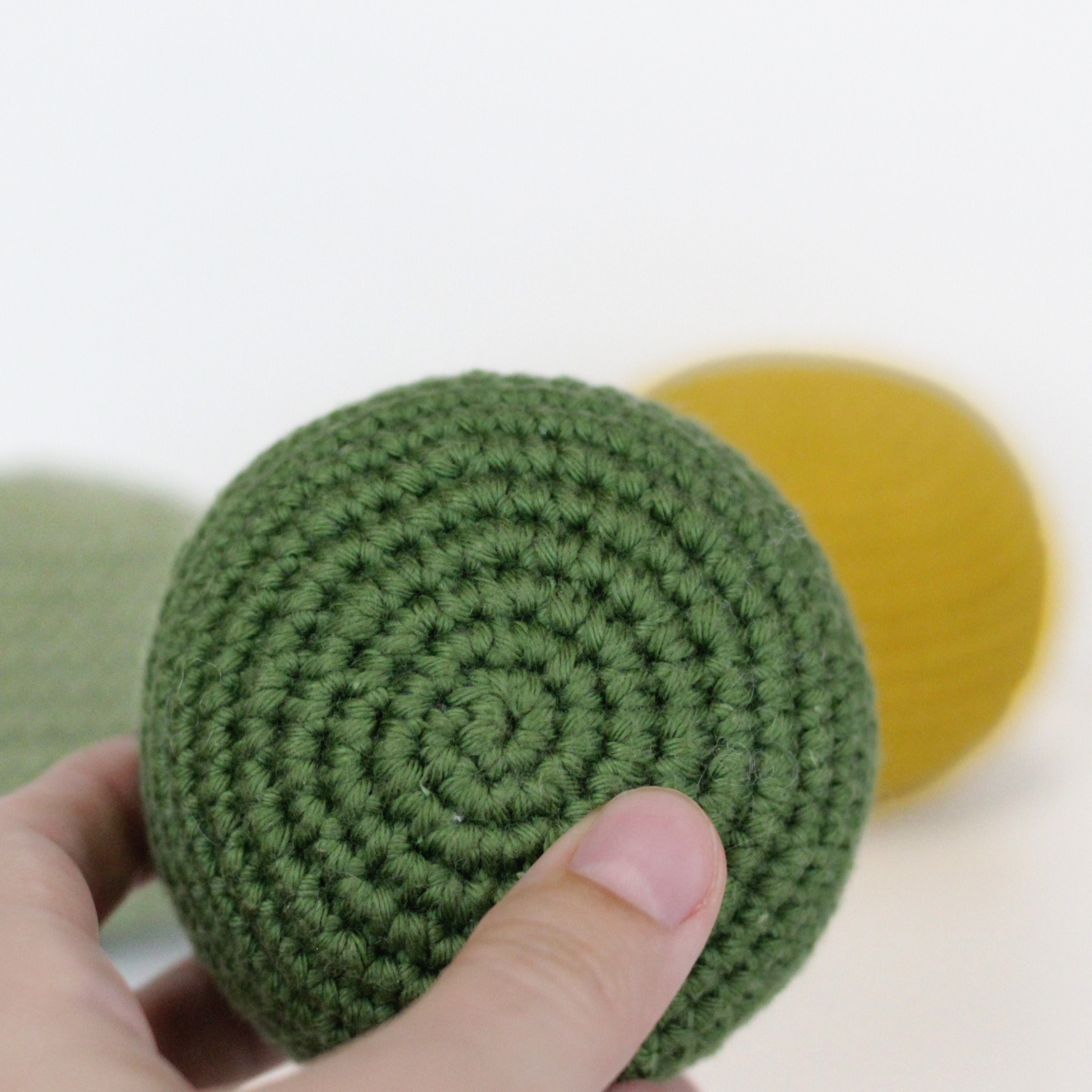 Forest Green crocheted ball held by a hand with a blurred yellow and moss green balls  on the background. Handmade crochet dog toys from Lana Royals Pet Wear.