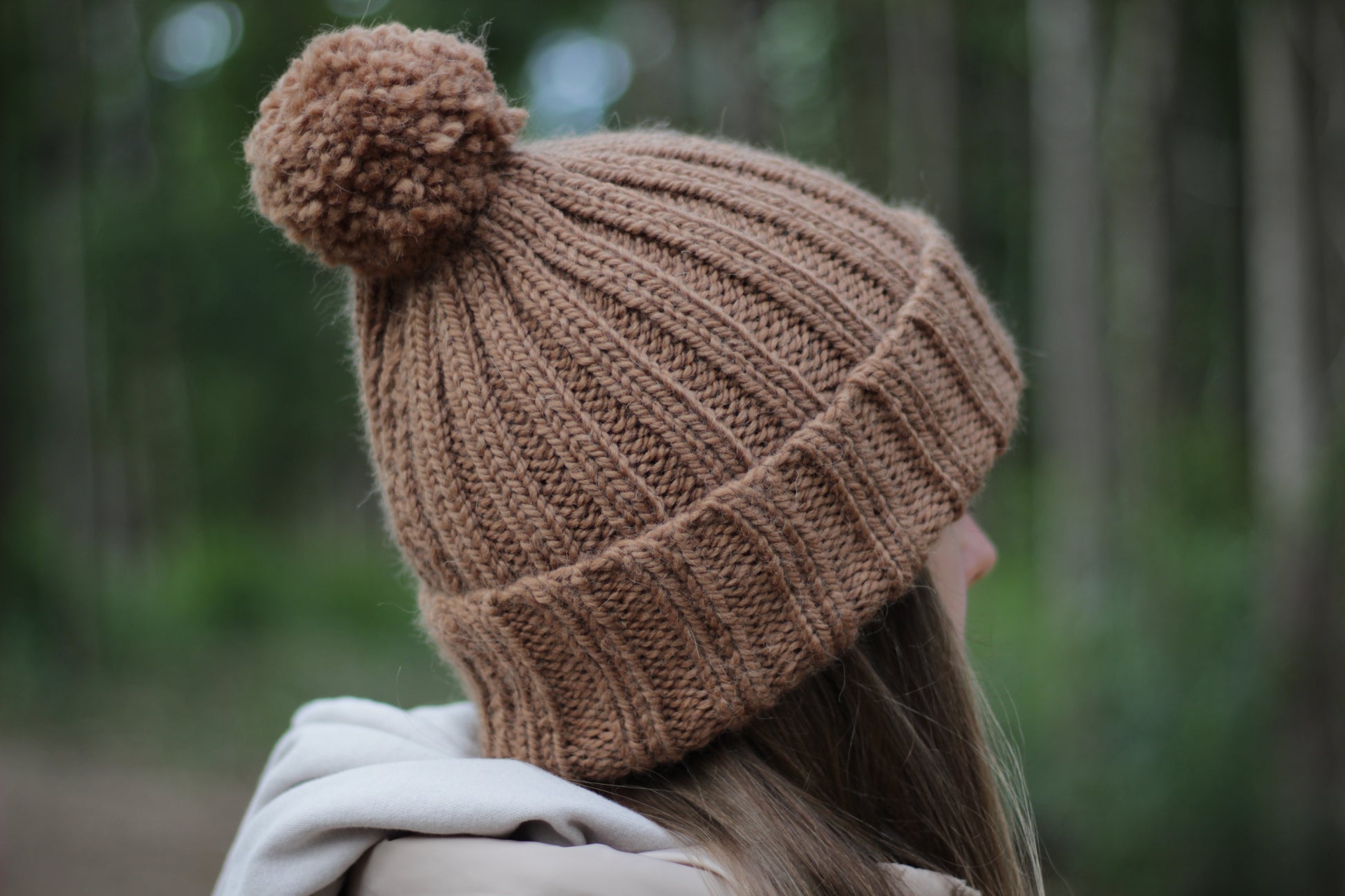 Person wearing a brown hand-knitted thick alpaca wool beanie with a pom-pom in a forest setting. Lana Royals Pet Wear Store.
