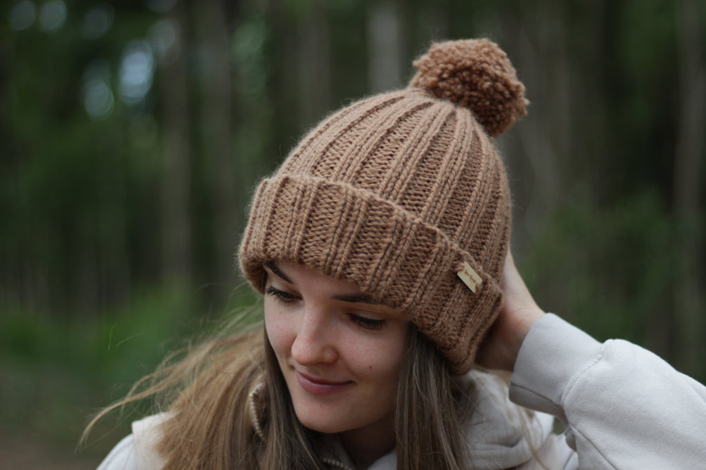 Woman wearing a brown hand-knitted thick alpaca wool beanie with a pom-pom in a forest setting. Lana Royals Pet Wear Store.
