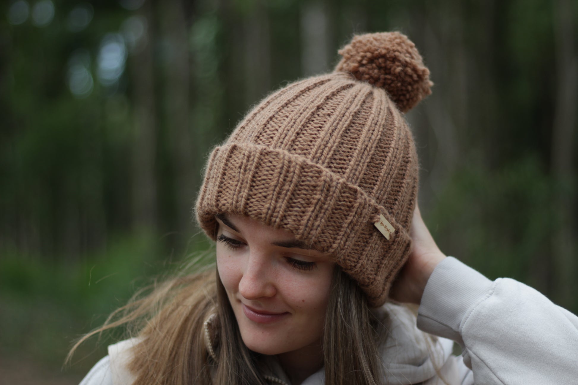 Woman wearing a brown hand-knitted thick alpaca wool beanie with a pom-pom in a forest setting. Lana Royals Pet Wear Store.