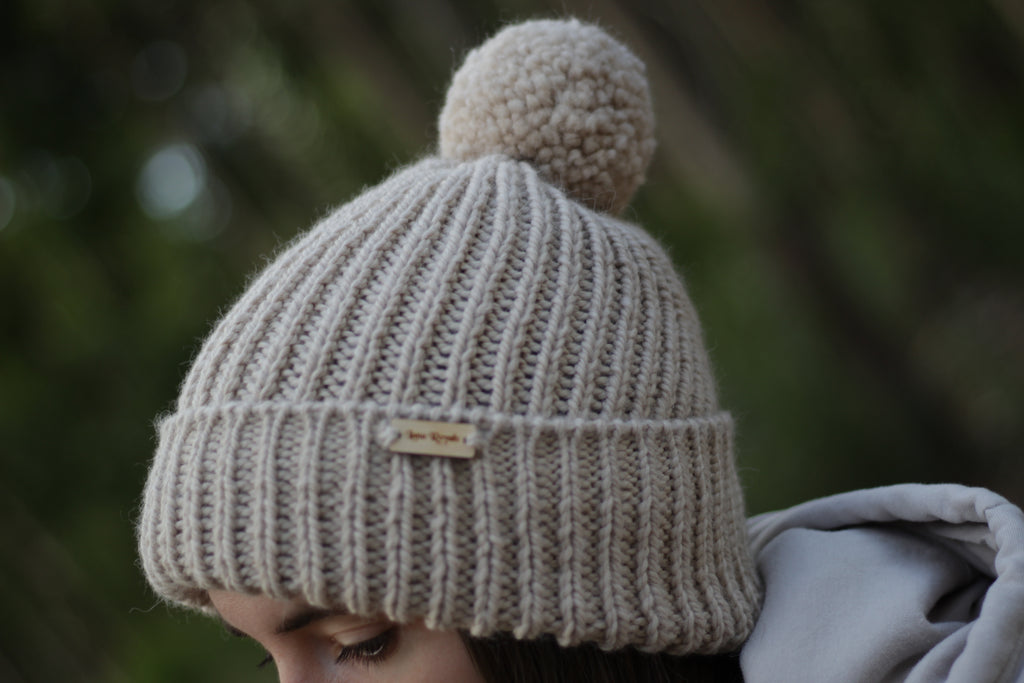 Beige alpaca wool knitted beanie with pom-pom on a blurred natural background. Handmade by Lana Royals Pet Wear Store.