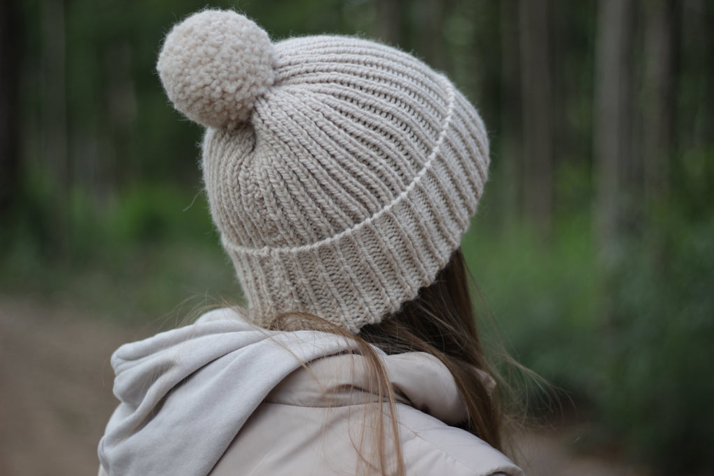 Person wearing a beige wool knit beanie with a pom-pom in a forest setting. Handmade by Lana Royals Pet Wear Store.